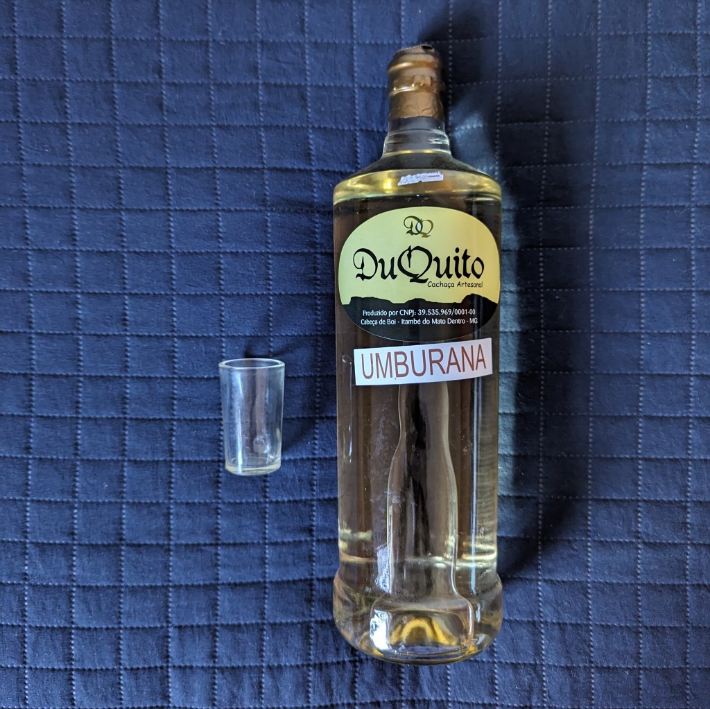 Do lado direito, garrafa de pinga Du Quito; do lado esquerdo, um copo pequeno de licor