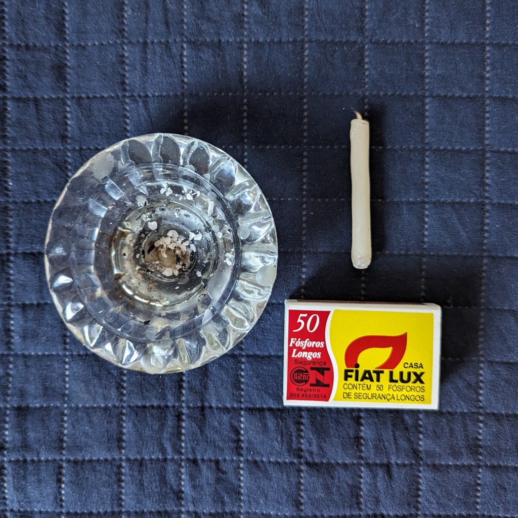 Castiçal de vidro do lado esquerdo, vela fina e caixa de fósforos do lado direito